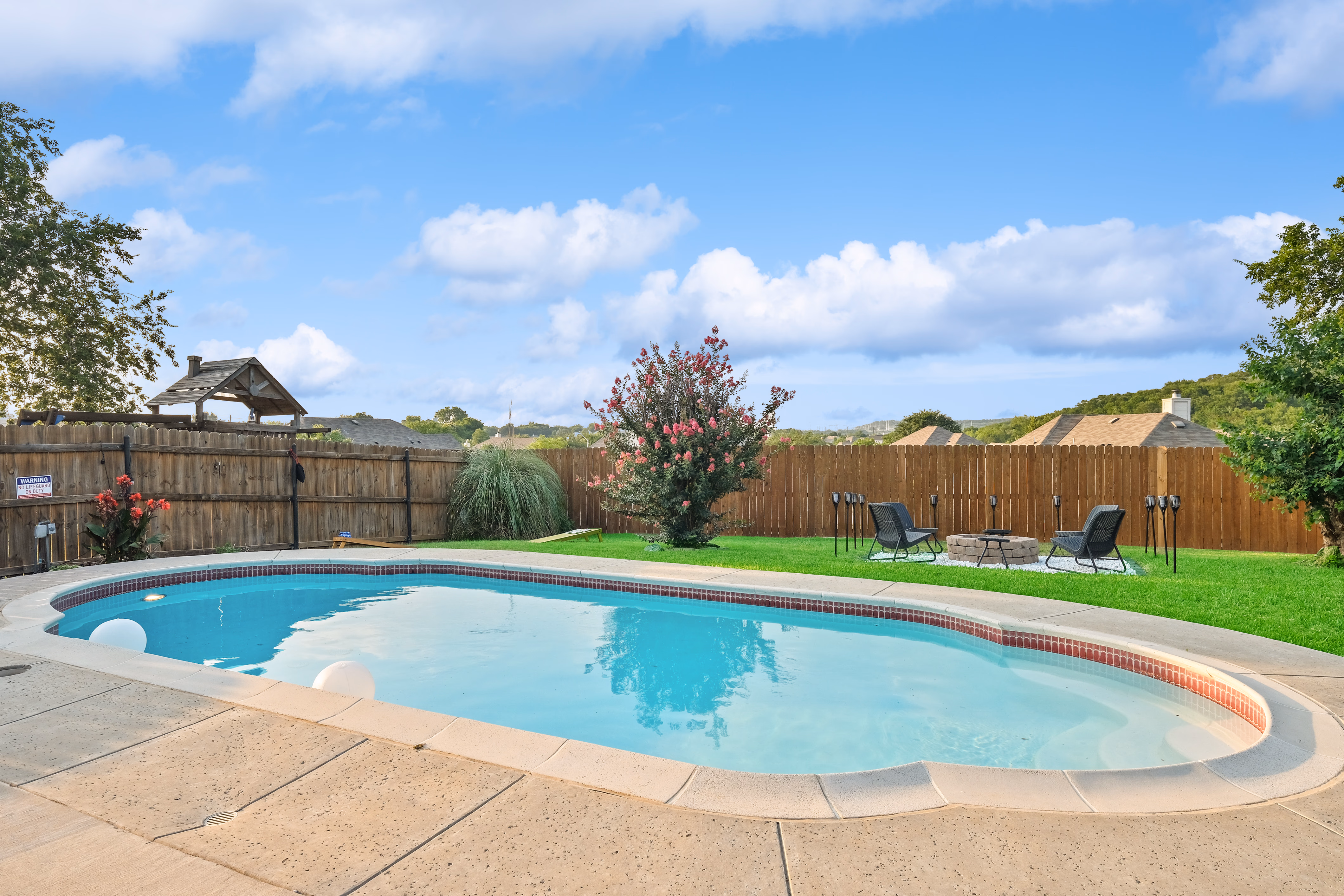 Pool with chairs and firepit table in fenced yard.